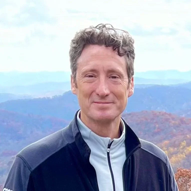 A man with curly brown hair dressed in a white and black jacket, standing outdoors with rolling hills and trees in the background.