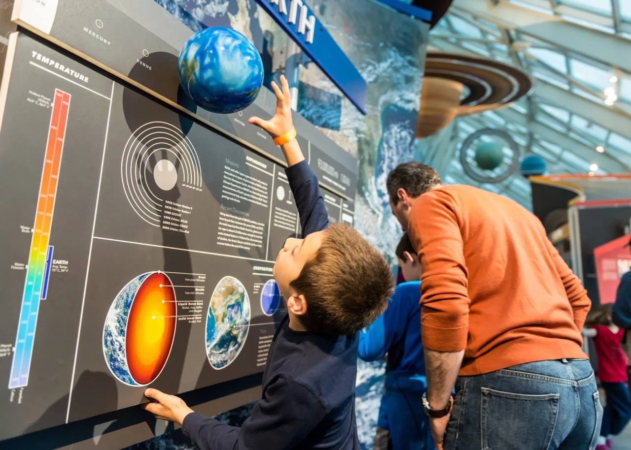 A boy reaching out to touch a model of the Earth in an educational exhibit about space and Earth's interior, with other visitors and space-themed displays in the background.