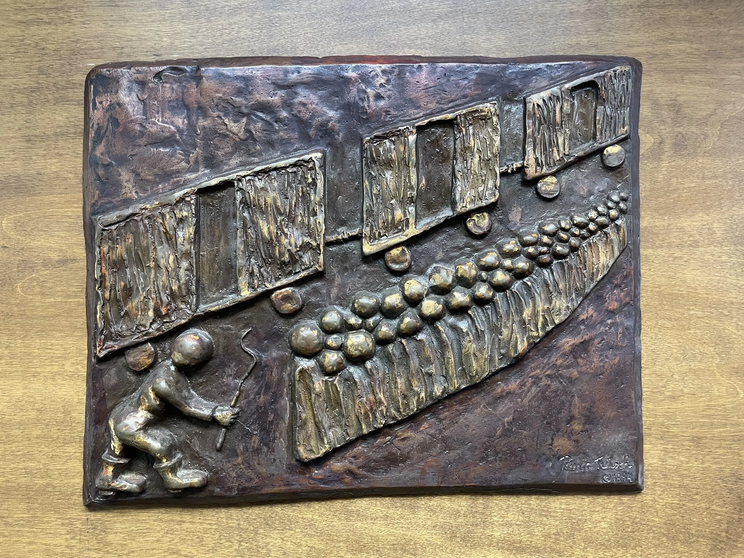 A metallic bronze-colored sculpture depicting a musical scene with a sheet of music, a trumpet, and several musical notes, set on a wooden surface.