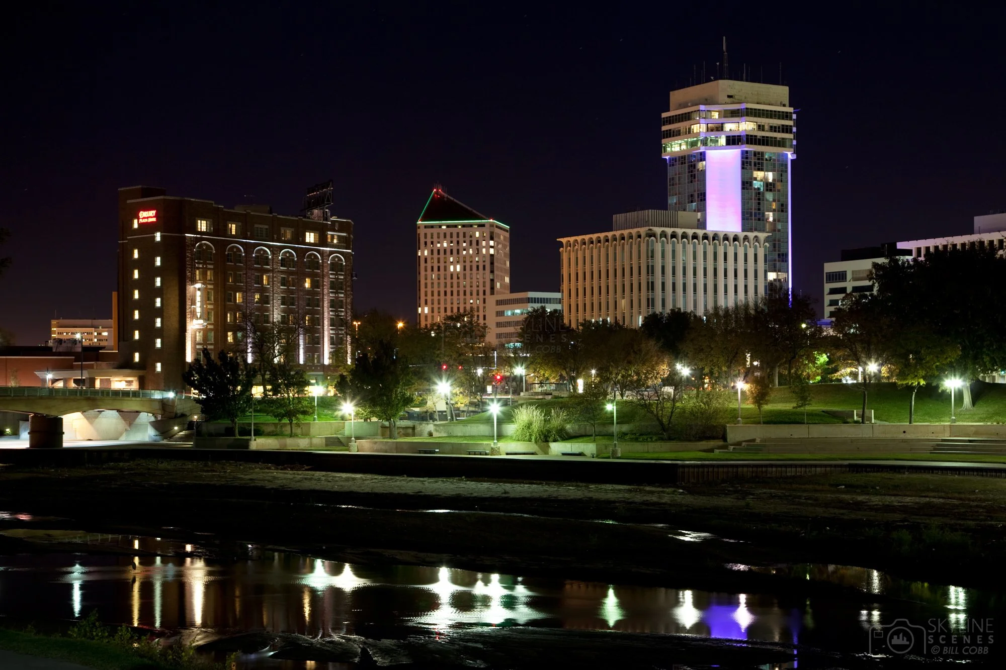 Nighttime cityscape with illuminated buildings reflecting on a river, trees, and park in the foreground.