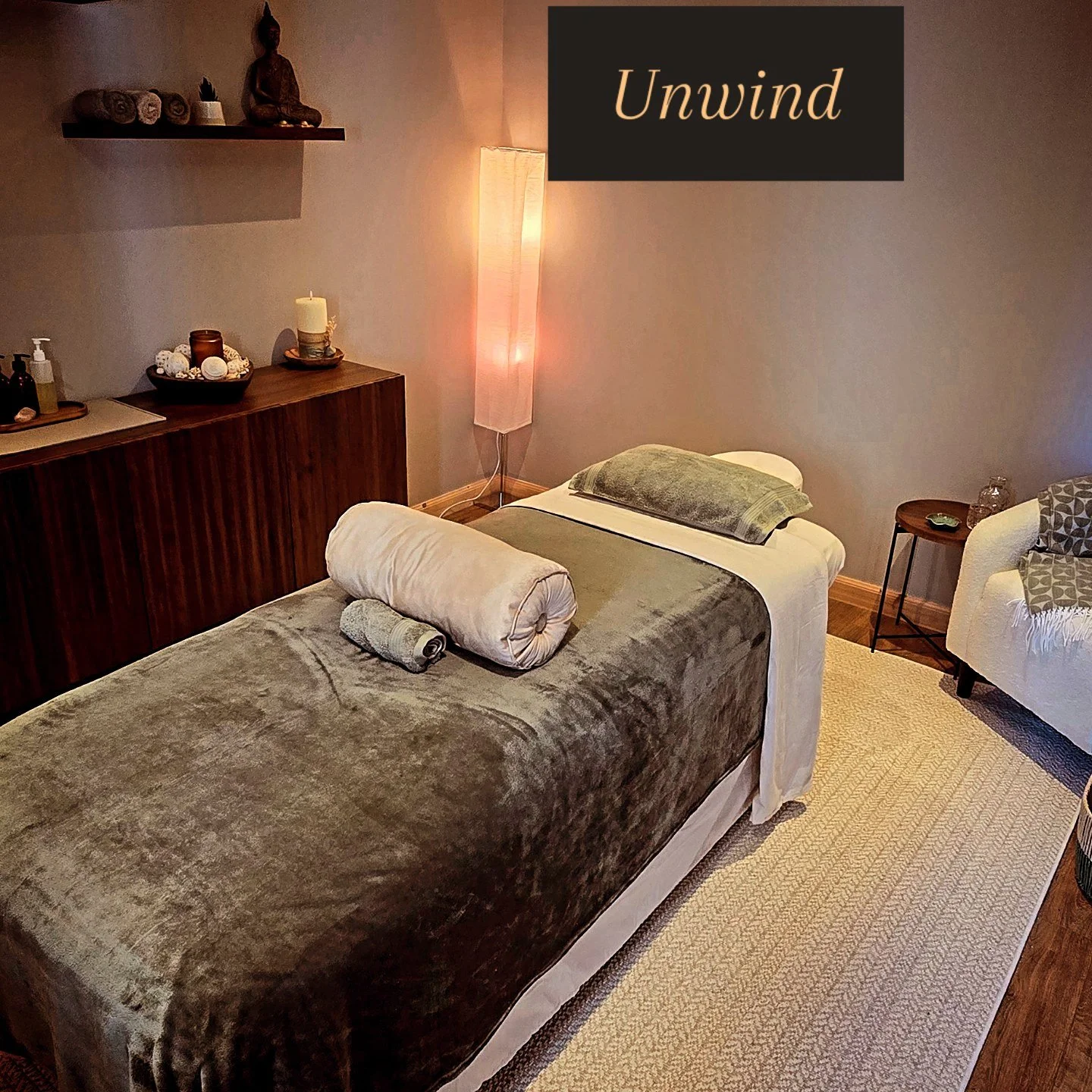 A massage room with a massage table covered in a plush brown blanket, with pillows and rolled towels on top. The walls are decorated with a floating shelf holding towels and a Buddha statue. There's a tall floor lamp emitting soft light, a small wooden side table with a few items, a white upholstered chair with a patterned throw blanket, and a beige rug on the wooden floor. A sign on the wall reads "Unwind."