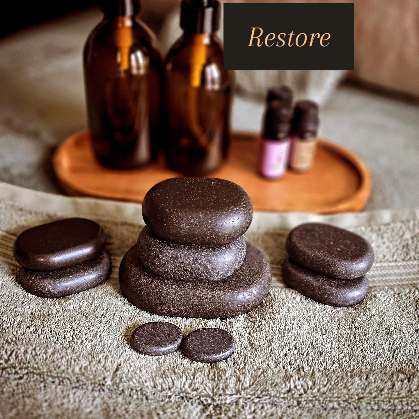 Stacked dark stones and smaller stones on a textured fabric, with massage oils and bottles in the background and a black box with the word 'Restore' on it.