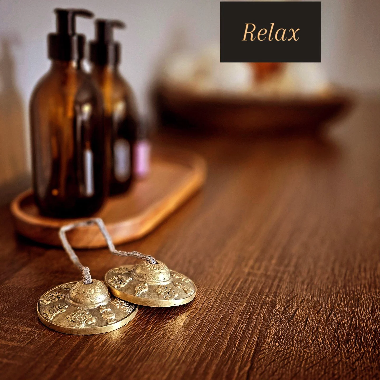 Close-up of a wooden table with metallic Tibetan prayer bells in the foreground and amber glass bottles on a tray in the background, with a black box containing the word 'Relax' in cursive font.