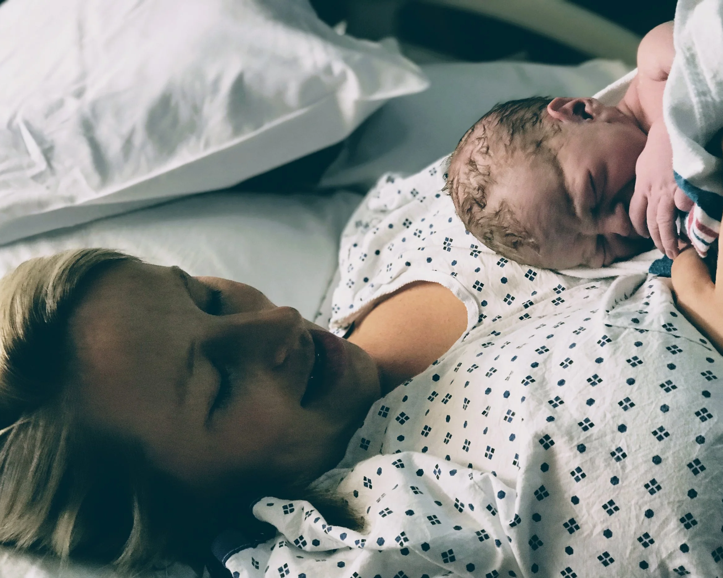 A newborn baby lying on a hospital bed, partially covered with a white blanket with blue patterns, with their head on a pillow and an adult woman with blonde hair looking at the baby.