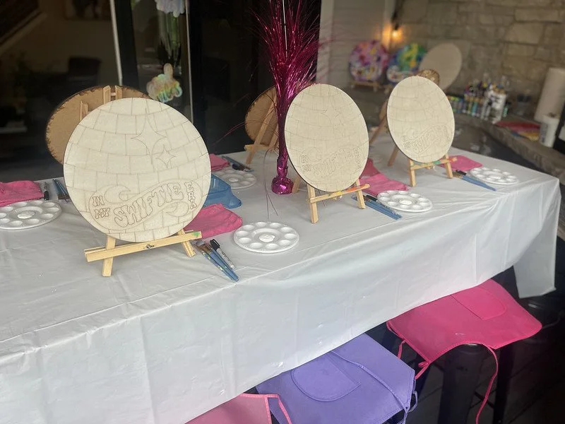 Paint canvases on easels set up on a table for an art activity or class, with paint palettes, brushes, and pink napkins. Future artwork seems to be designed for a celebration or event.