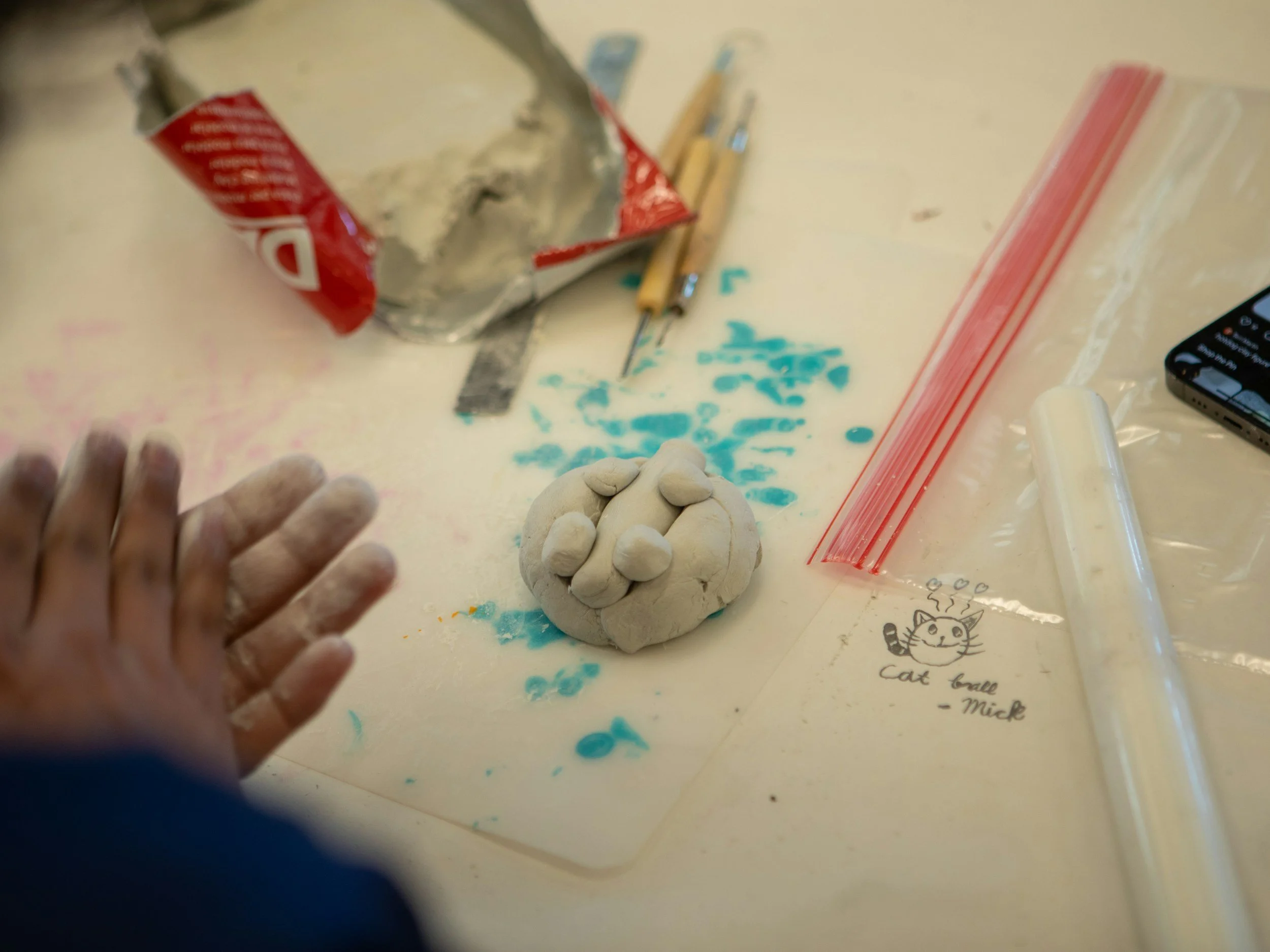 A workspace with a handful of dried clay balls, an open bag of flour, sculpting tools, a plastic bag with a red zipper, and a small digital device on a table marked with a cartoon cat drawing and the words 'cat ball - Mick'.