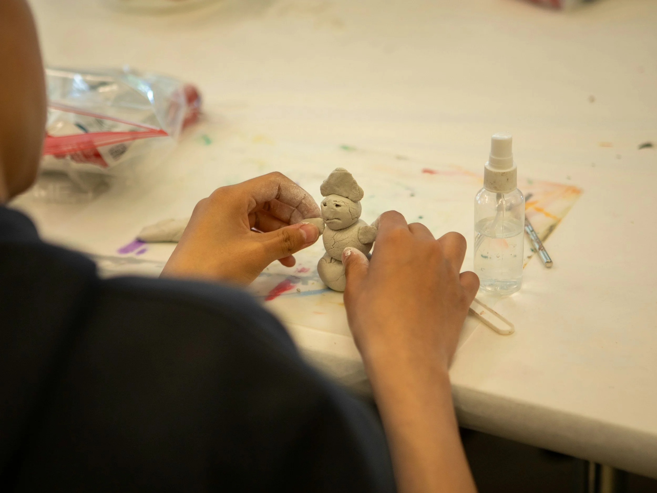 A person making a small clay figure of a person with a hat with their hands on a colorful, paint-splattered table, with a spray bottle and art supplies nearby.
