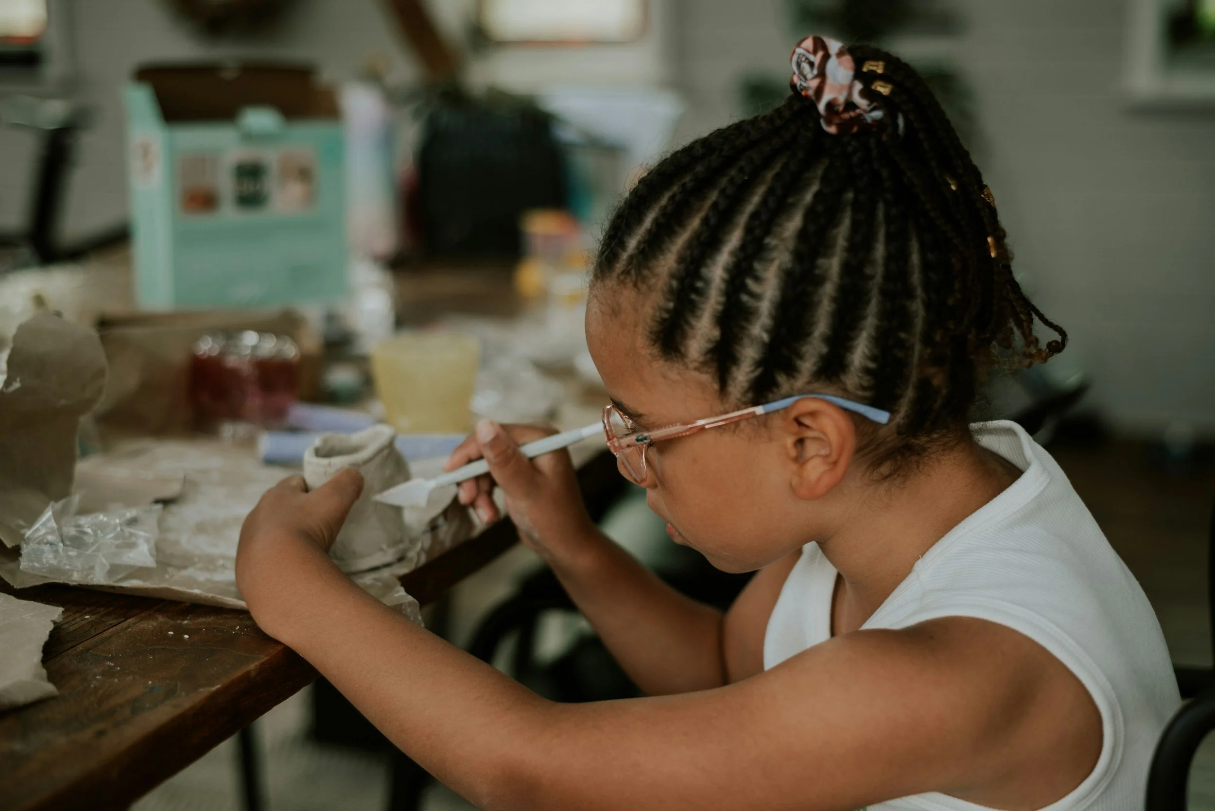 A young girl with black braided hair and pink glasses sitting at a wooden table, painting or working on an art project with concentration, in a room with a cluttered background.