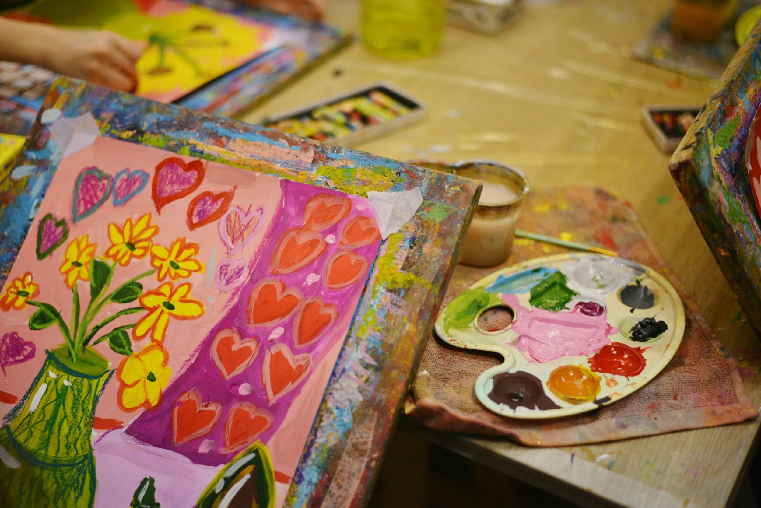 Paintings of hearts and flowers on a canvas, with art supplies and a paint palette nearby in an art studio.