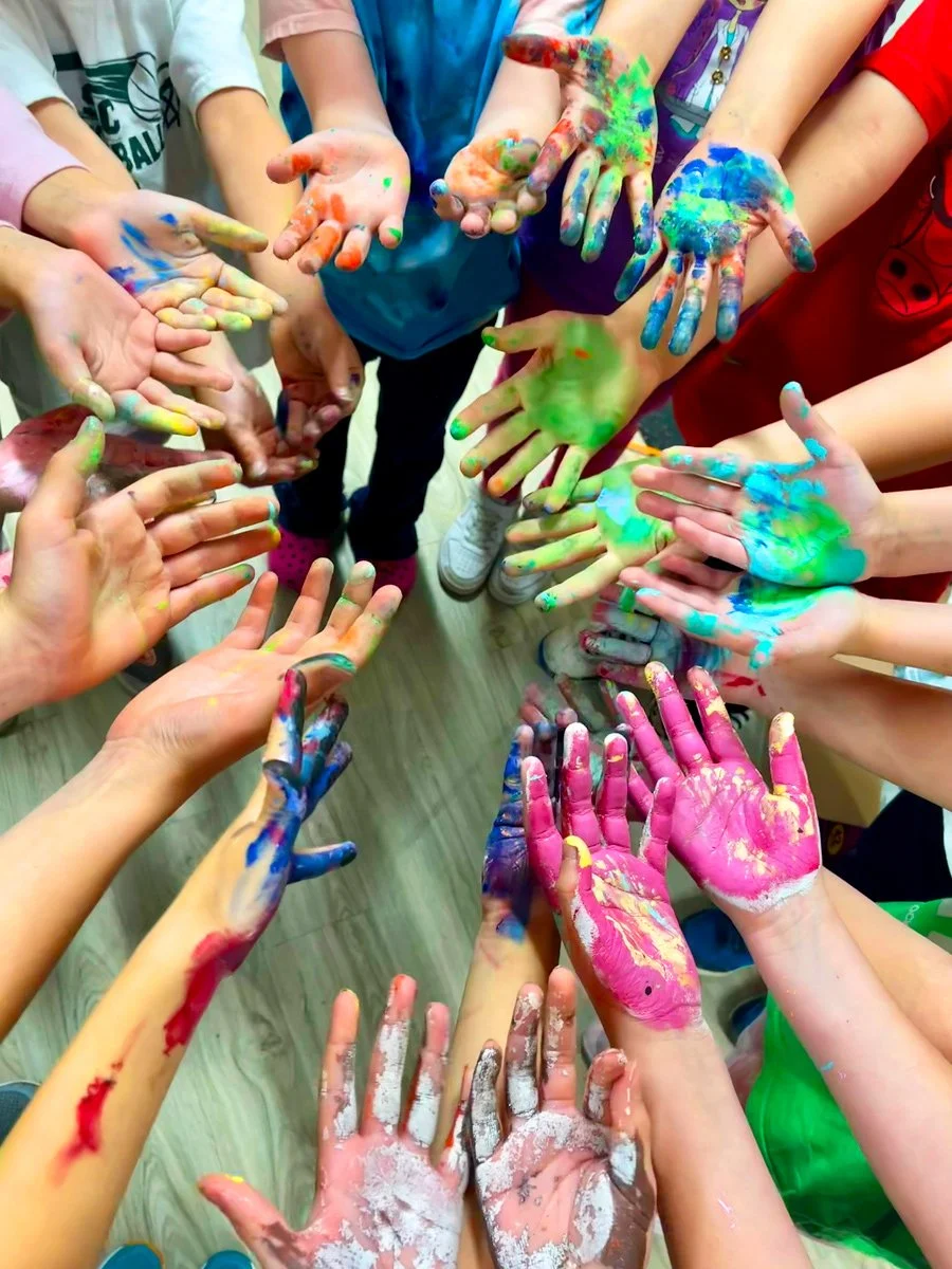 Group of children with colorful paint-splattered hands reaching towards the camera in a circle.