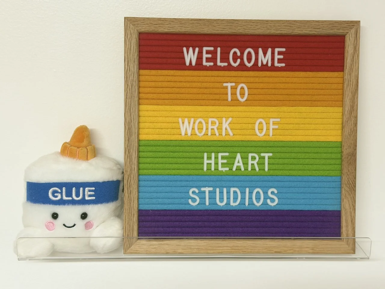 Colorful framed sign reading, 'Welcome to Work of Heart Studios,' with rainbow-striped background in red, orange, yellow, green, blue, and purple, next to a plush toy of a smiling glue bottle with a happy face, rosy cheeks, and an orange cap.