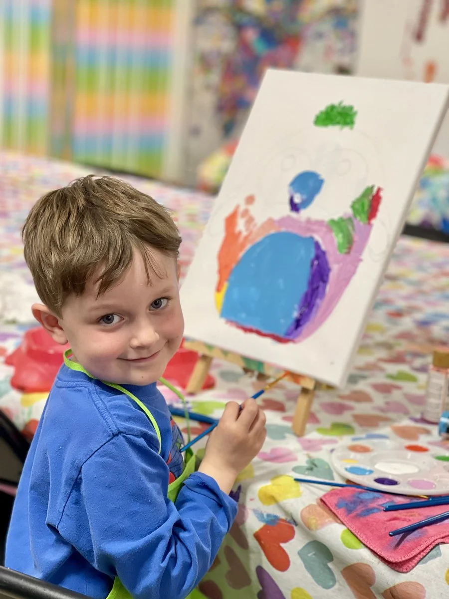 A young boy with light brown hair wearing a blue shirt and a green apron smiles while painting on a canvas at an art station. The canvas displays colorful abstract shapes. The background shows a vibrant, colorful display of art supplies and artwork.