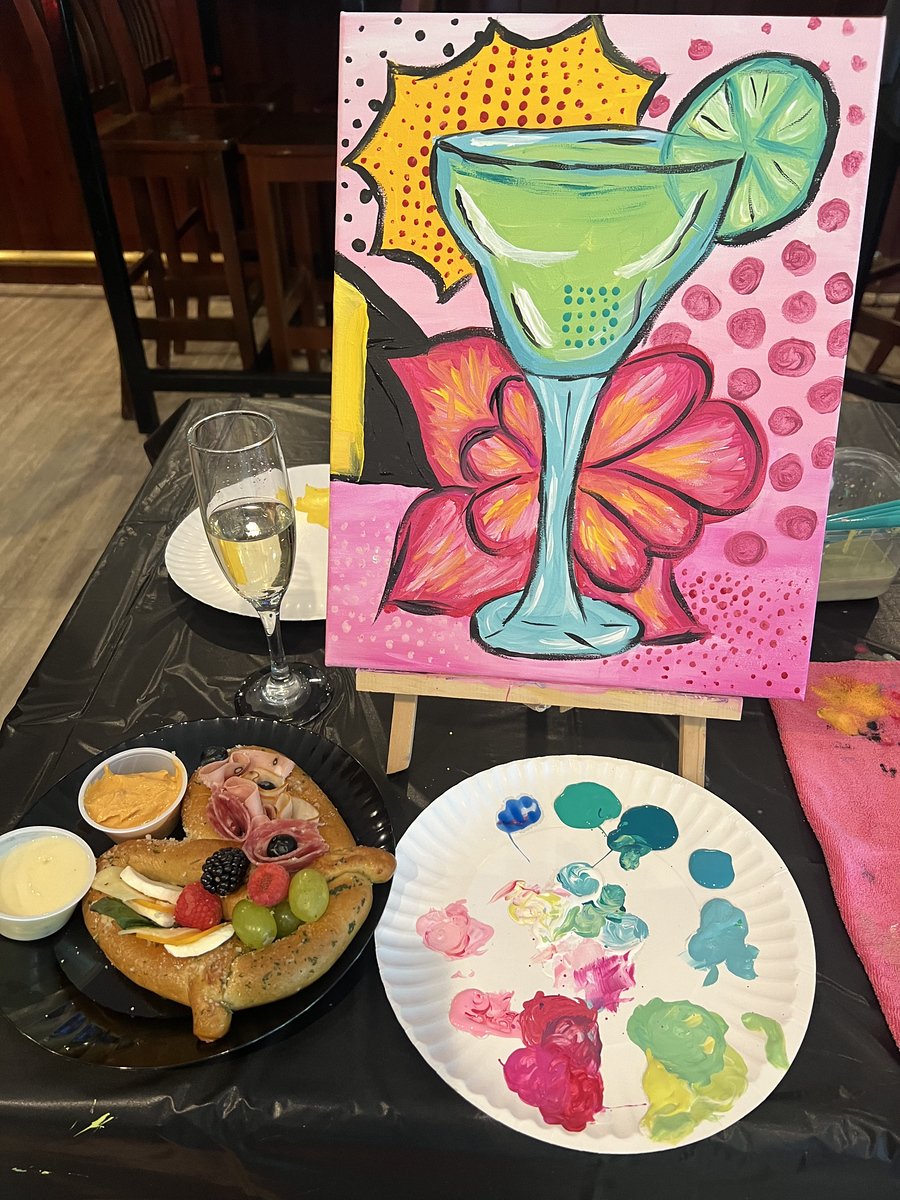 Colorful painting of a green margarita with a lime slice on the rim, set against a pink background with pink dots and a yellow burst design. In front of the painting is a plate with a cooked chicken, decorated with grapes, berries, cheese, and meats, along with containers of dip or sauce. There is also a glass of champagne and a paper plate with paint splashes on the table.