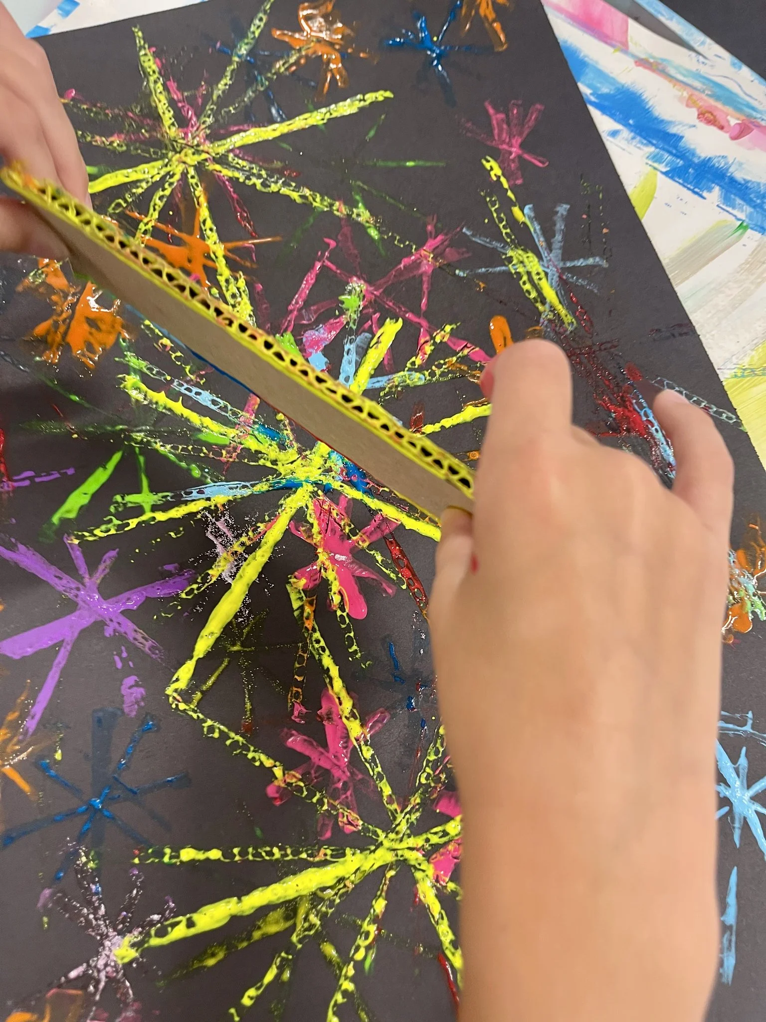 Child creating a colorful explosion of paint on black paper using sticks dipped in vibrant paint colors.