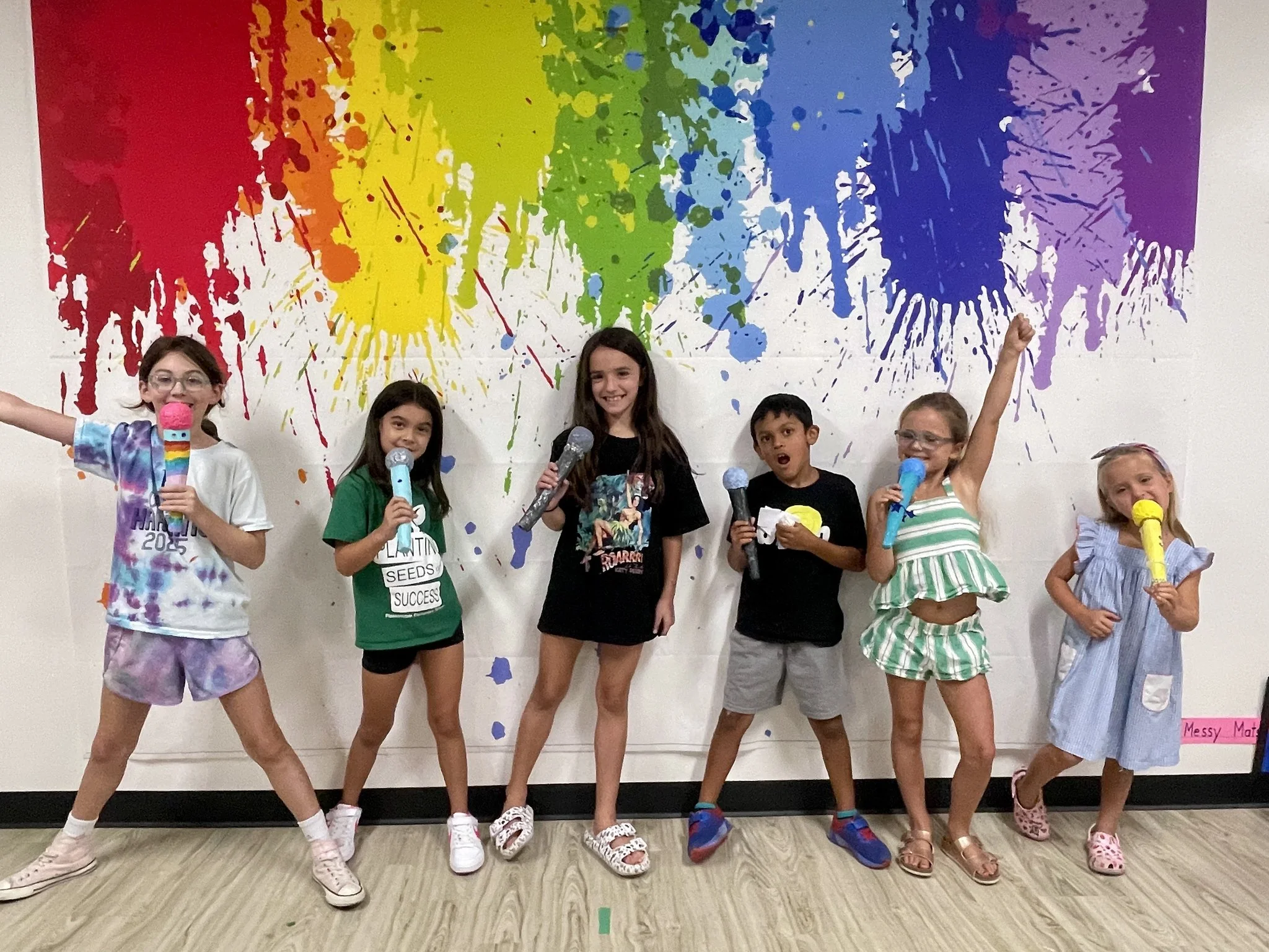 A group of six children standing in front of a multicolored paint splatter wall. They are holding toy microphones, smiling, and posing for the camera. The children are wearing casual summer clothes and some are wearing glasses.