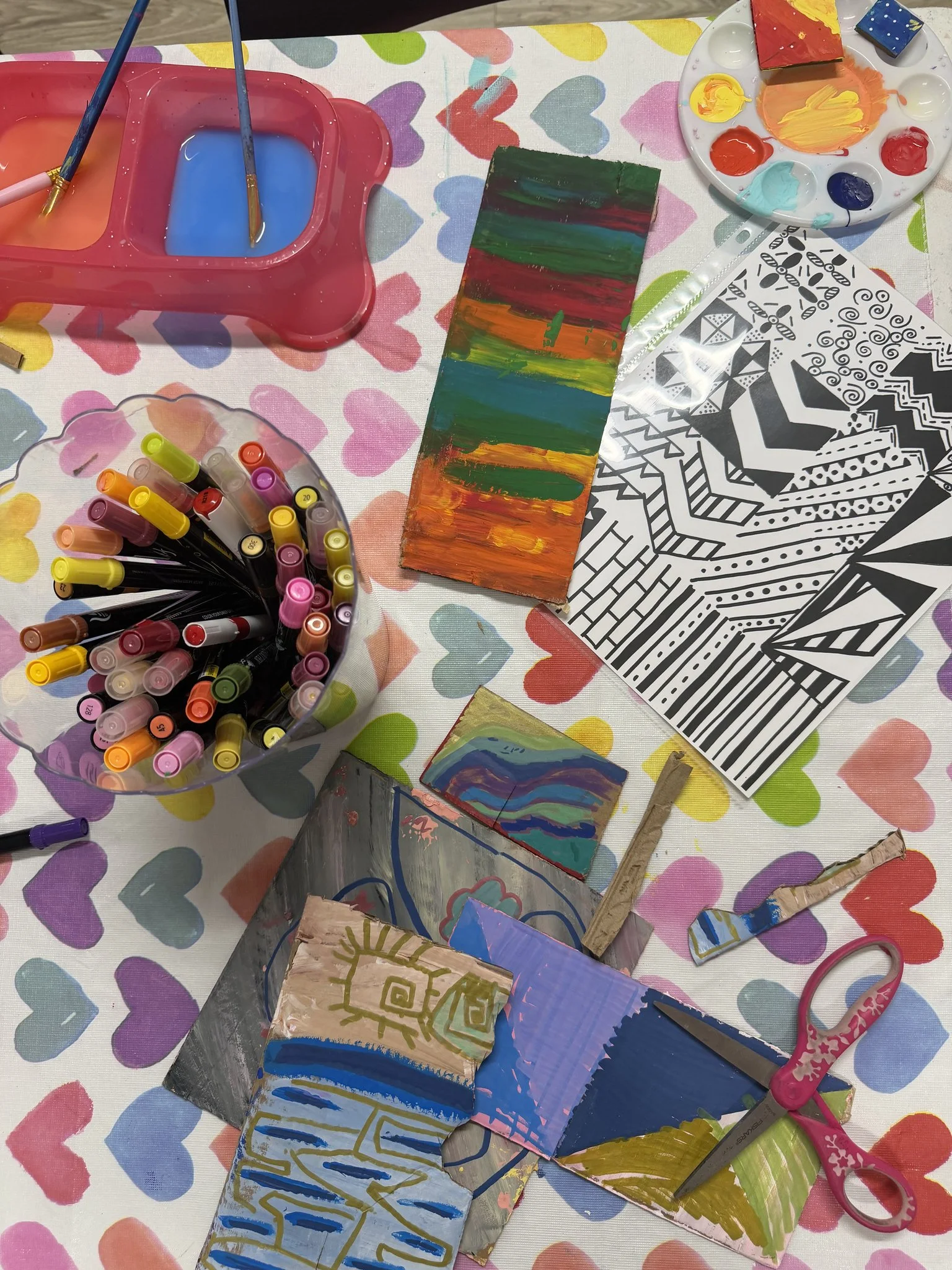 Art supplies including paintbrushes, paint palettes, markers, and painted rocks on a colorful heart-patterned tablecloth.