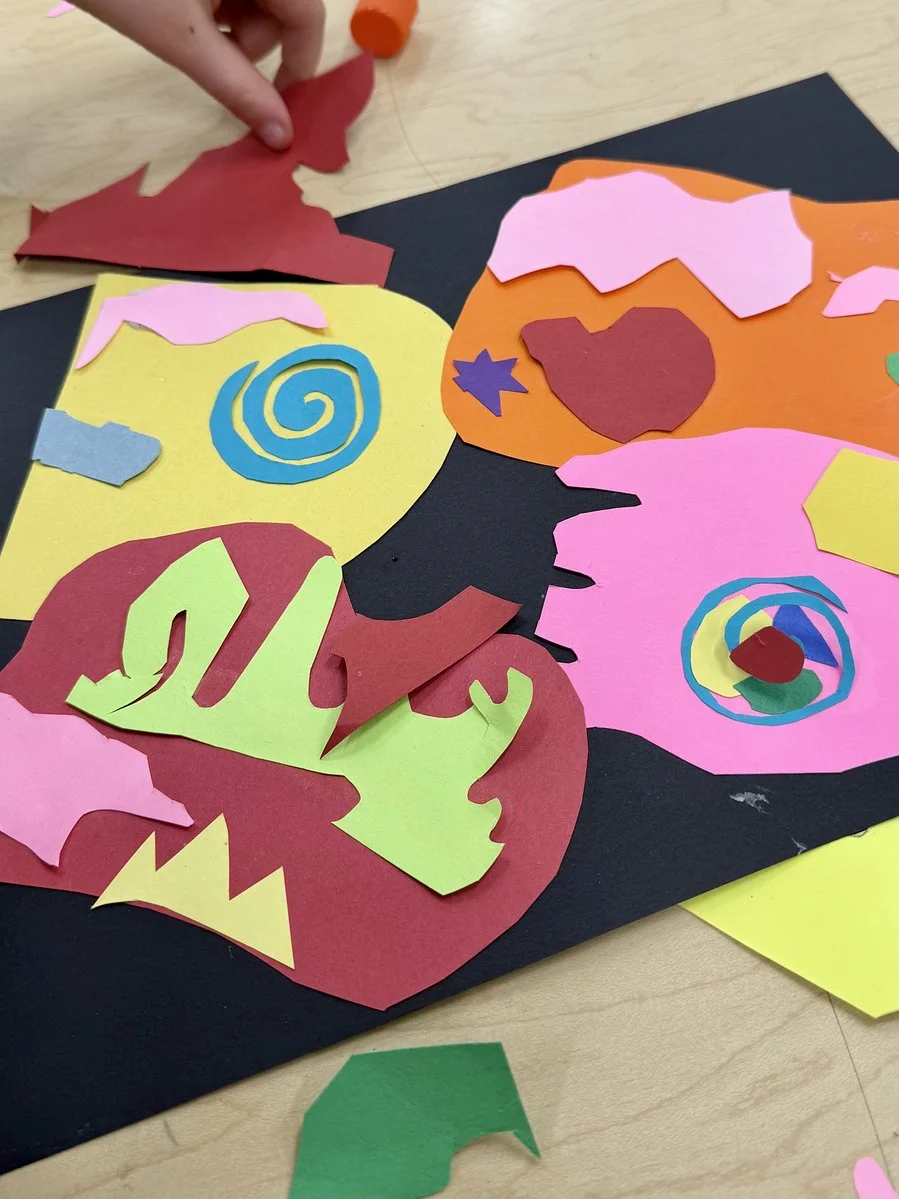 Colorful paper cutouts of hearts, stars, and swirling patterns on black paper, with a person's hand adjusting a red paper shape. There are additional cutouts on a light-colored wooden table.