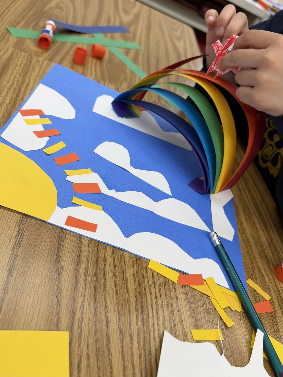 Child is creating a rainbow craft with colored paper strips, gluing them onto a blue paper background with cloud shapes and a yellow sun, using scissors and glue on a wooden table.