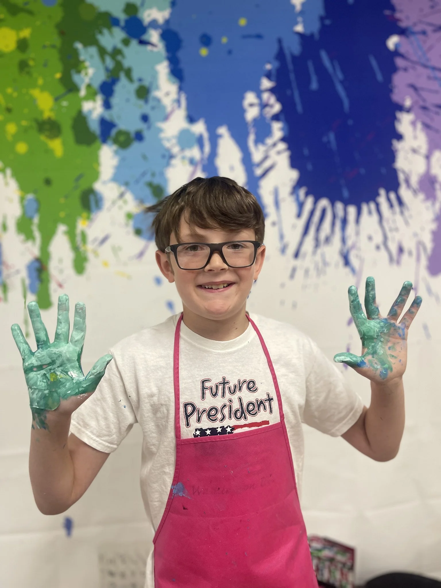 A boy wearing glasses and a pink apron with the words "Future President" on it, smiling and showing painted hands raised in the air, standing in front of a colorful wall with paint splatters.
