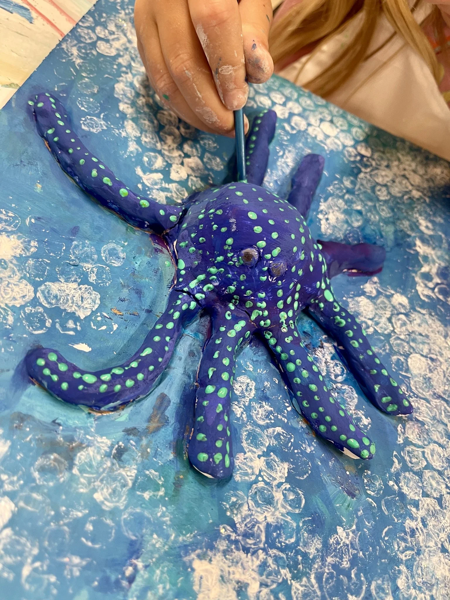 A person painting a large, detailed octopus sculpture with purple and green accents. The person's hand, covered in white paint, is holding a small brush, working on the sculpture's head. The scene is set on a textured blue surface.