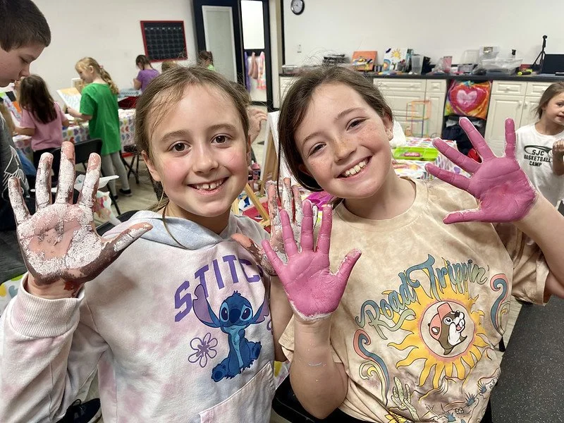 Two smiling girls with painted pink hands showing art project in a classroom.