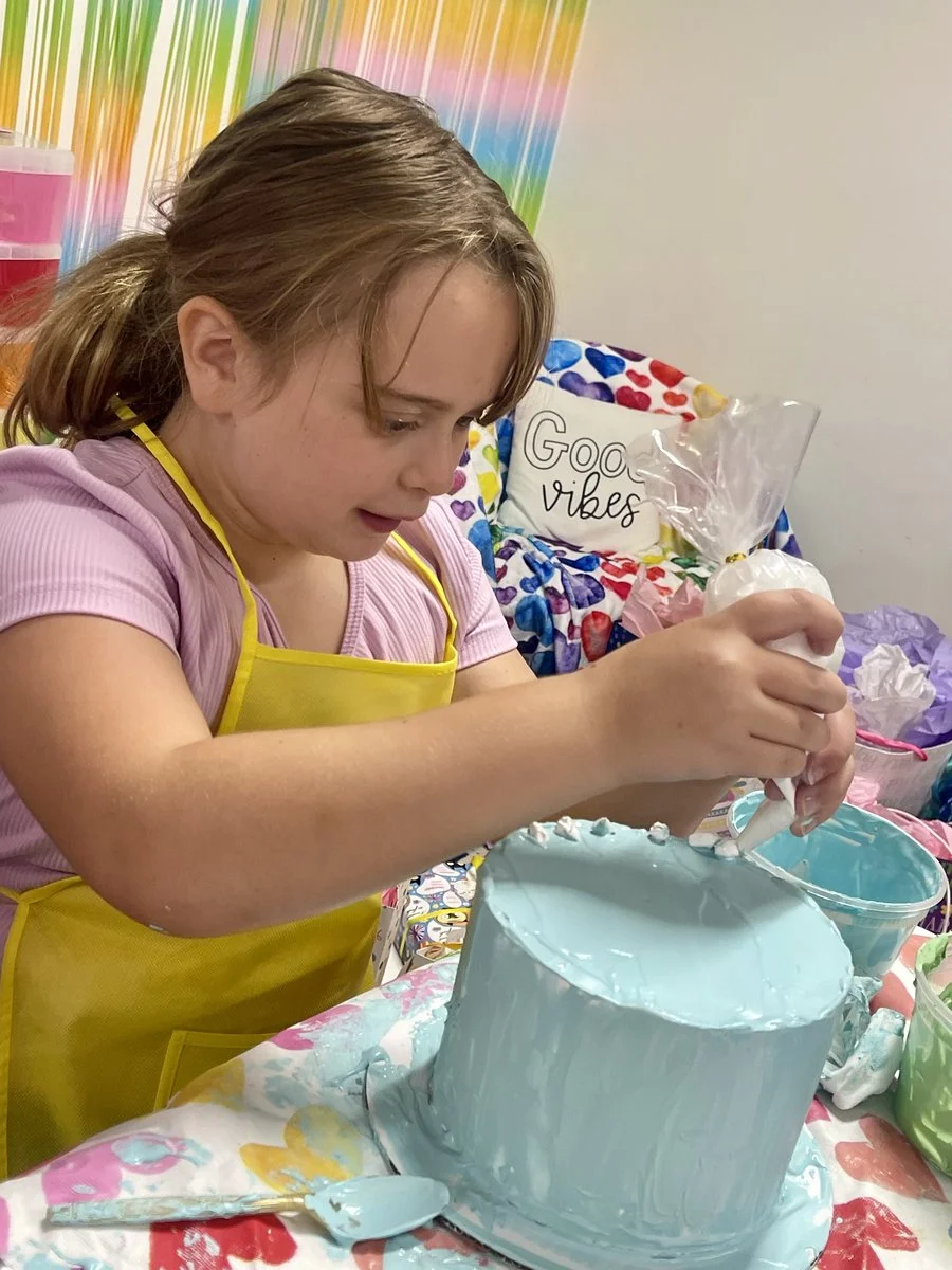 A young girl wearing a pink shirt and a yellow apron is decorating a cake with blue frosting. She is piping icing onto the cake in a bright room decorated with colorful balloons and a pillow that says 'Good vibes'.