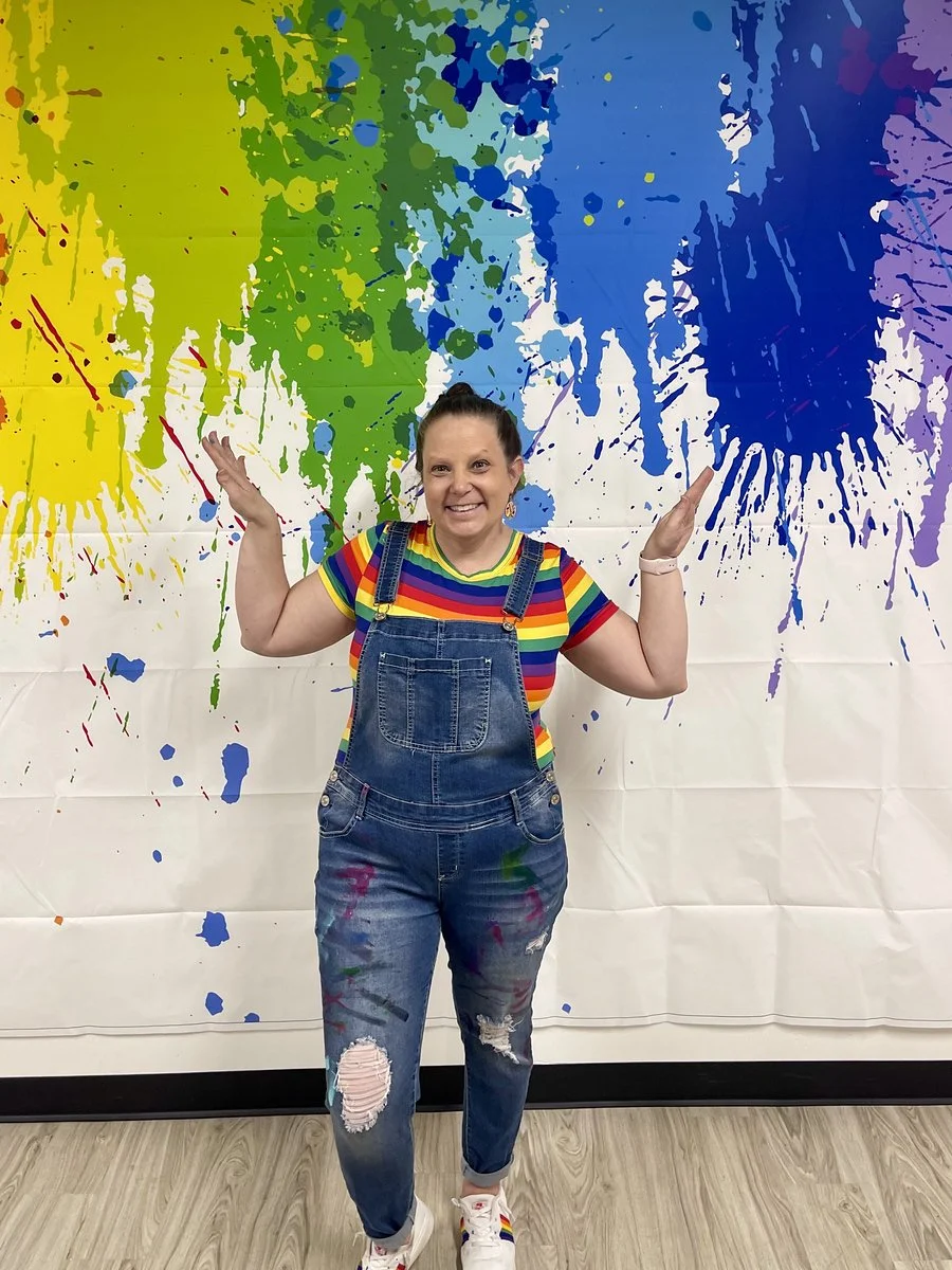 Woman smiling and raising her arms in front of a colorful splatter paint backdrop. She wears rainbow-striped shirt, denim overalls, and sneakers.