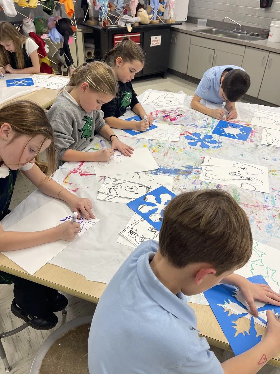 Six children are sitting around a table, drawing and coloring winter and holiday-themed pictures on paper, with art supplies scattered across the table in an arts classroom.
