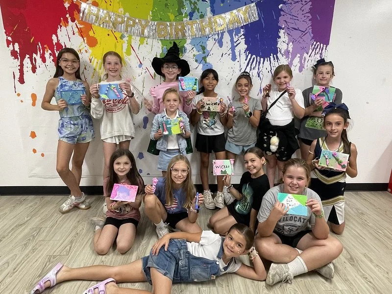 Group of young girls celebrating a birthday, holding colorful handmade cards and gifts, standing in front of a vibrant rainbow paint splatter wall with a "Happy Birthday" banner.