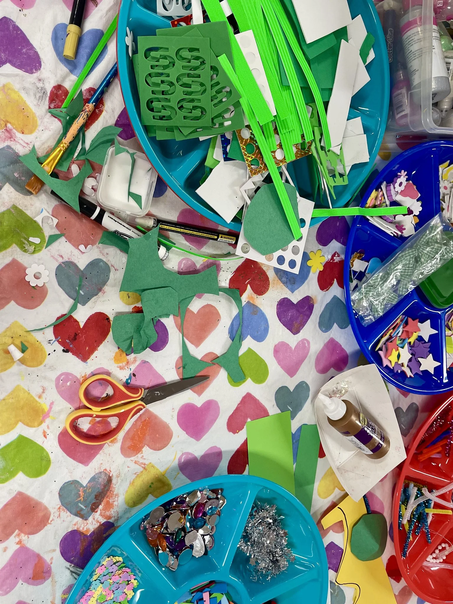 A colorful table filled with craft supplies including green foam sheets, scissors, glue, rhinestones, sequins, and various small craft embellishments, some on plastic trays.