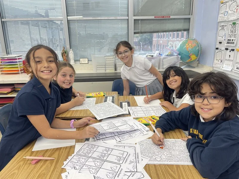 Five children sitting at a table in a classroom, working on coloring and drawing activities. There are worksheets and coloring supplies on the table, with a globe and educational posters in the background.
