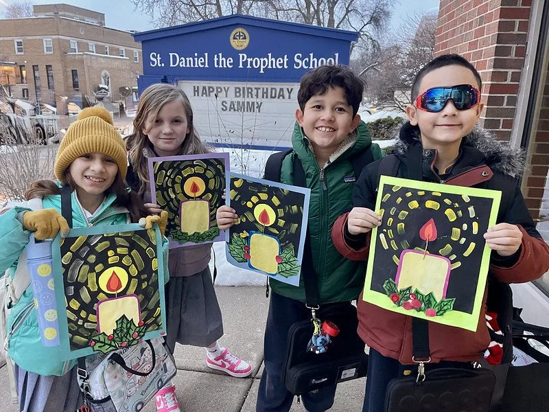 Four children standing outside in front of a sign that reads "St. Daniel the Prophet School," holding colorful drawings of a candle with a flame, surrounded by yellow and pink patterns, celebrating a holiday at school.