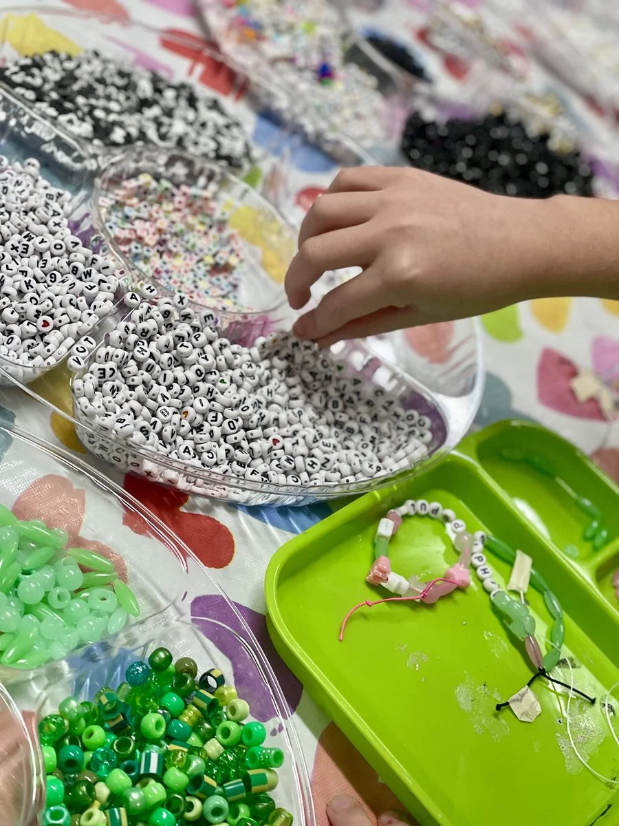 A person making jewelry using colorful beads from various bowls, with a focus on a hand reaching into a bowl of white letter beads.