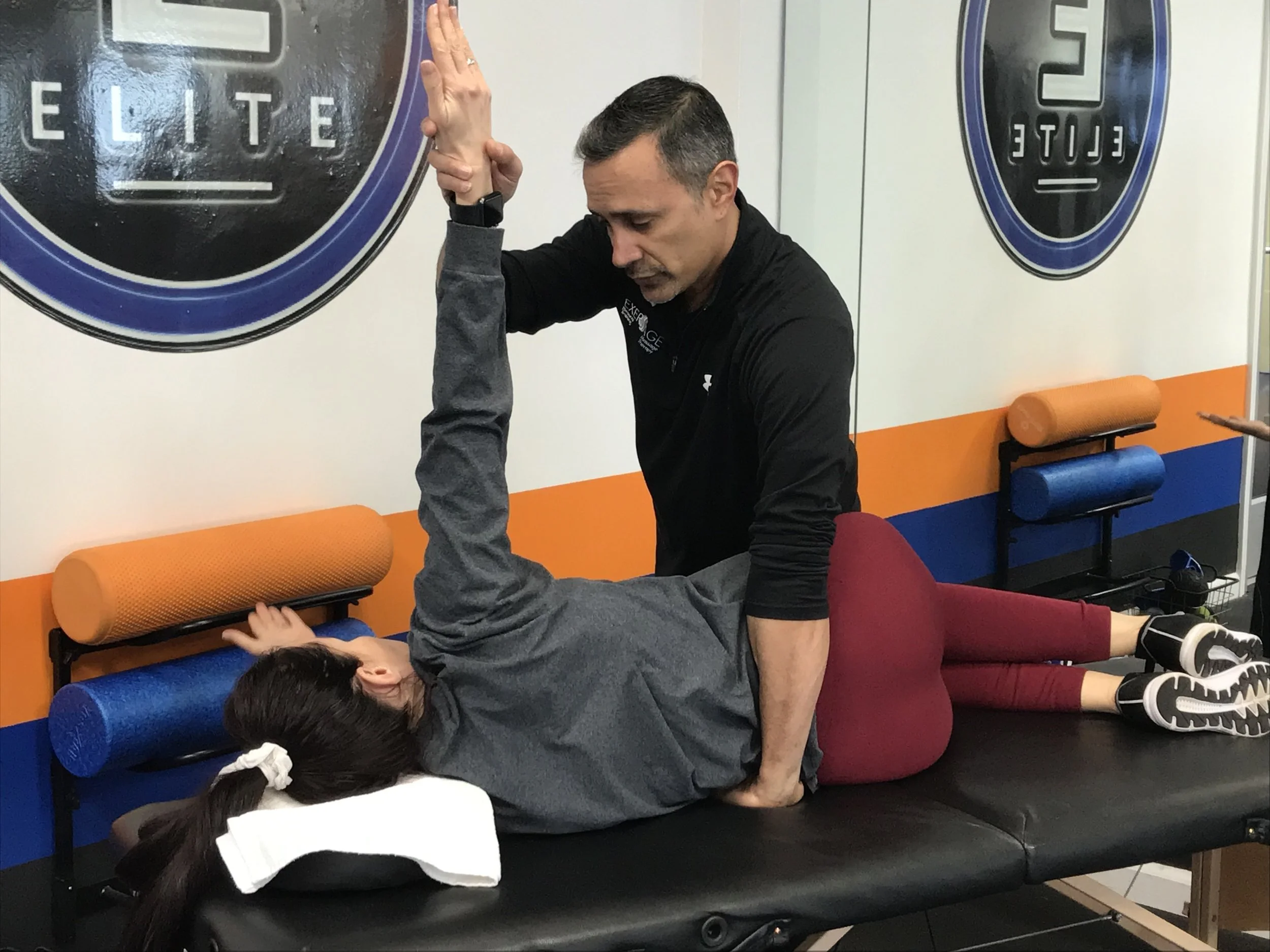 A man assisting a young girl doing a backbend exercise on a black padded bench in an exercise or therapy room. The girl is lying on her back with her legs supported by him, and she is reaching upward with her arms. The room has colorful foam rollers and mirrors on the wall.