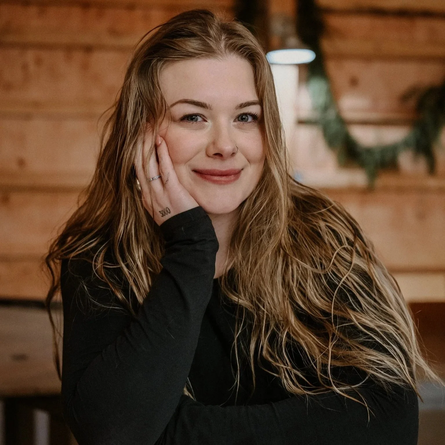 A young woman with wavy light blonde hair, fair skin, and a nose ring, smiling and resting her face on her hand, leaning on a bar, with a wooden background and greenery in the background.