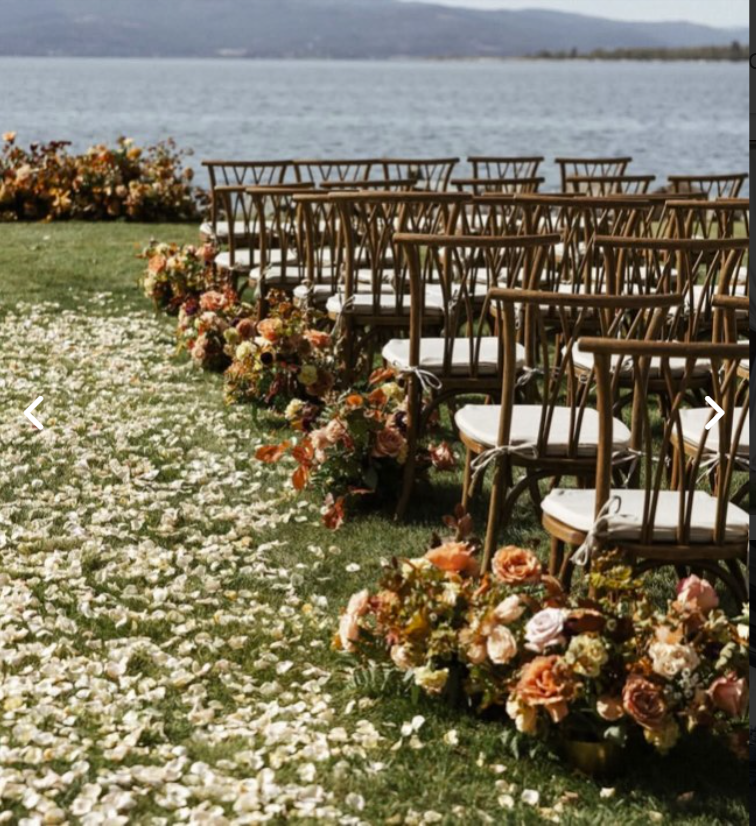 Outdoor wedding ceremony setup by the water with chairs, floral arrangements, and flower petal aisle.
