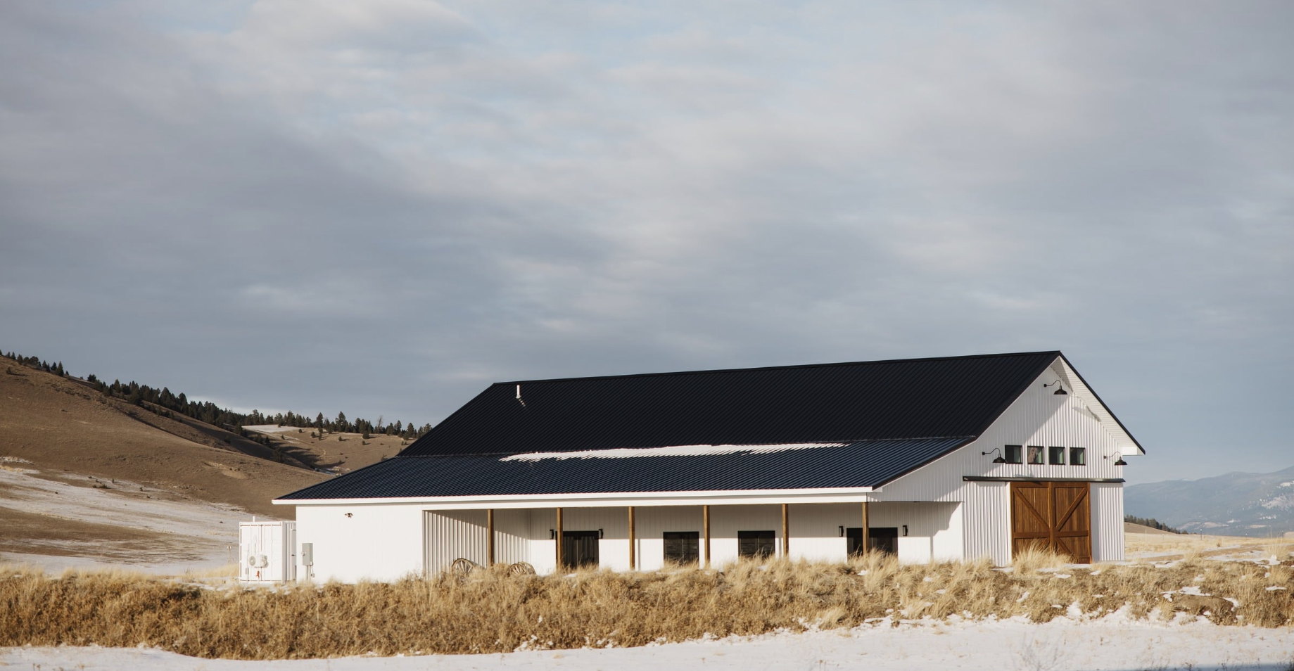 A large white barn with a black metal roof and a wooden sliding door, situated in a rural landscape with dry grass, snow patches, and rolling hills in the background under a cloudy sky.