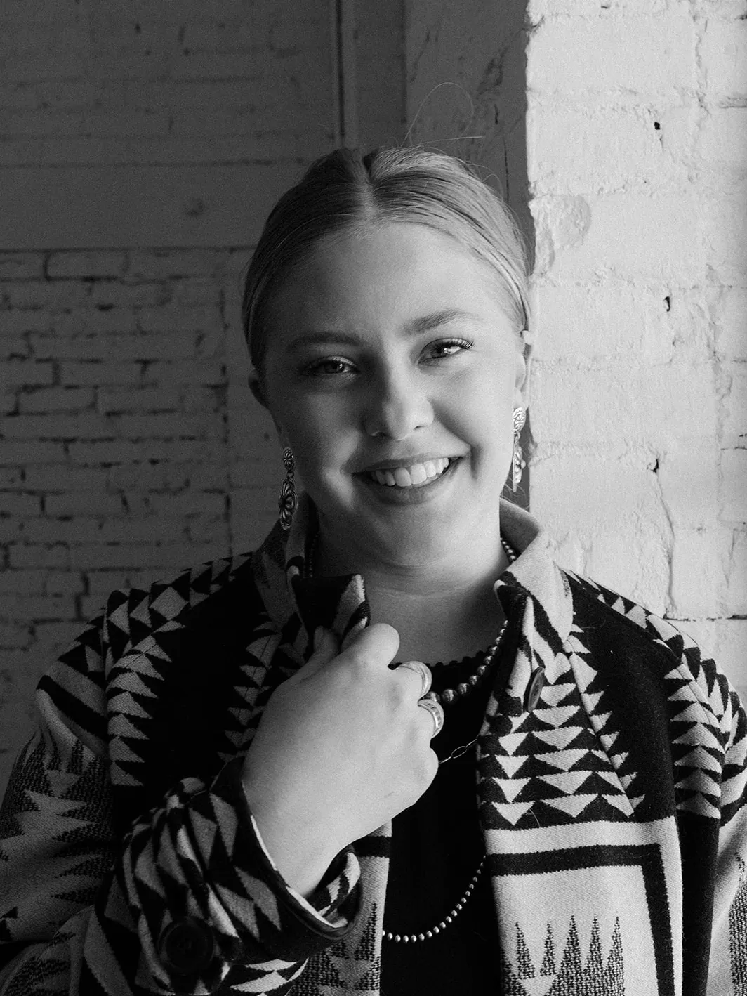 A woman with light hair pulled back, smiling, wearing earrings, necklaces, and a patterned jacket, standing against a brick wall.