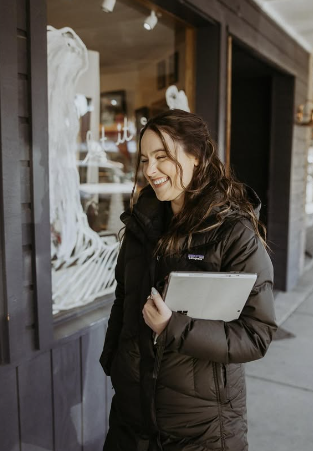 A woman smiling and walking outside while holding a laptop and some papers, wearing a black jacket.