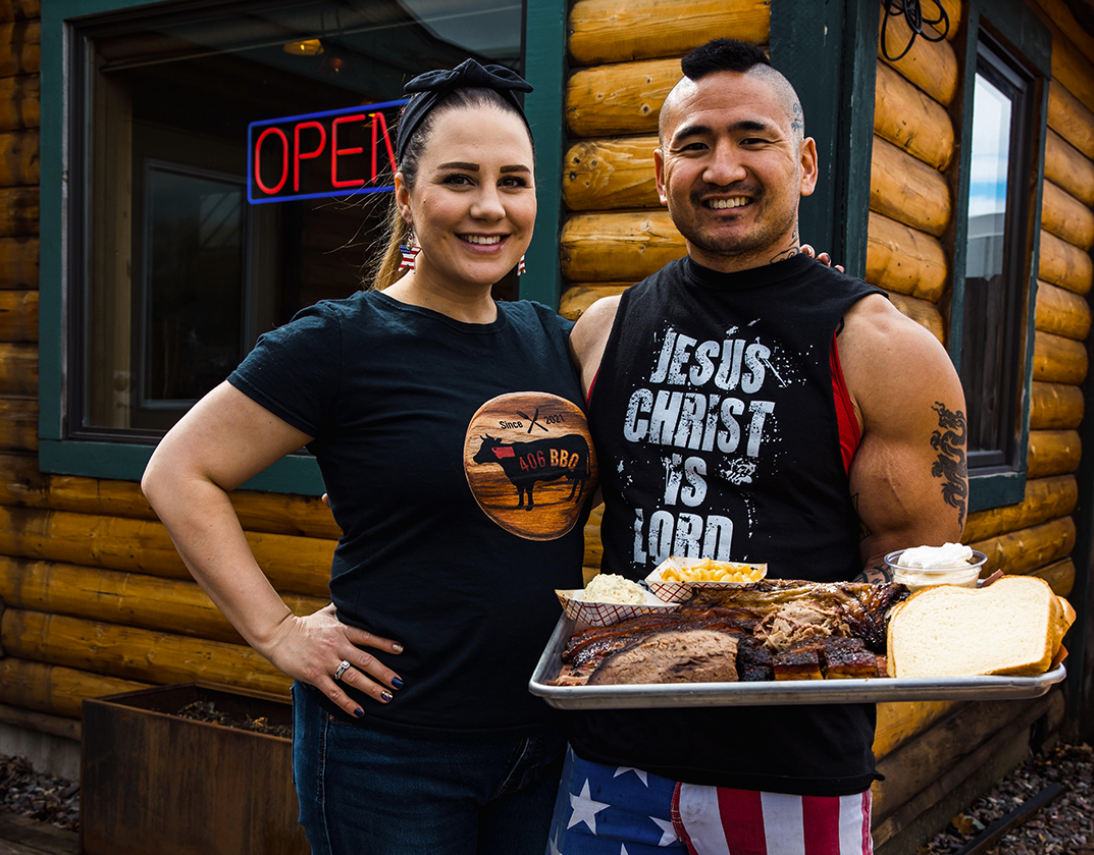 A woman and man smiling outside a log cabin restaurant. The woman is wearing a black t-shirt with a BBQ logo, and the man is wearing a black sleeveless shirt with religious text. He's holding a tray of various barbecue foods, including meat, bread, and sides.