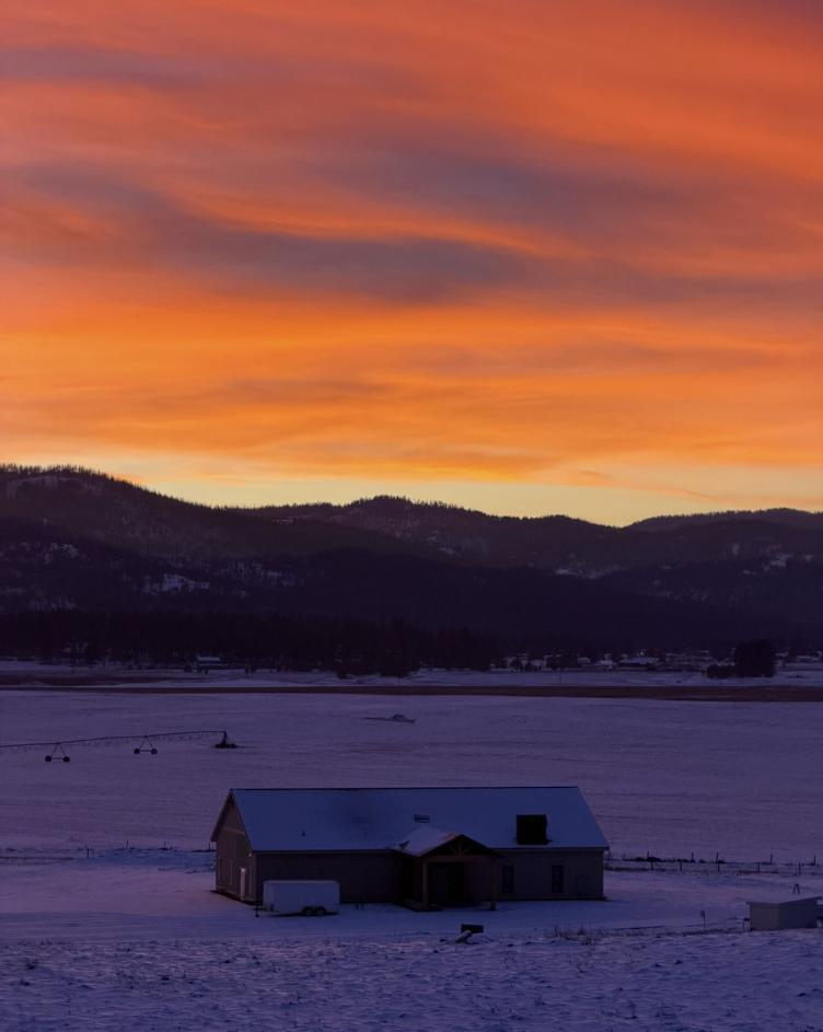 A snowy landscape at sunset with a house in the foreground and mountains in the background