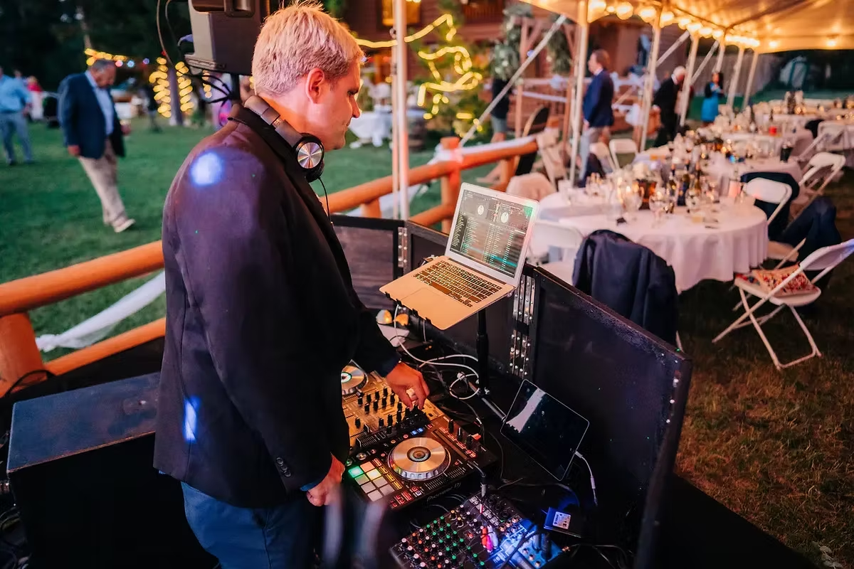 DJ in black suit playing music at nighttime event with tables, chairs, and string lights under tent.