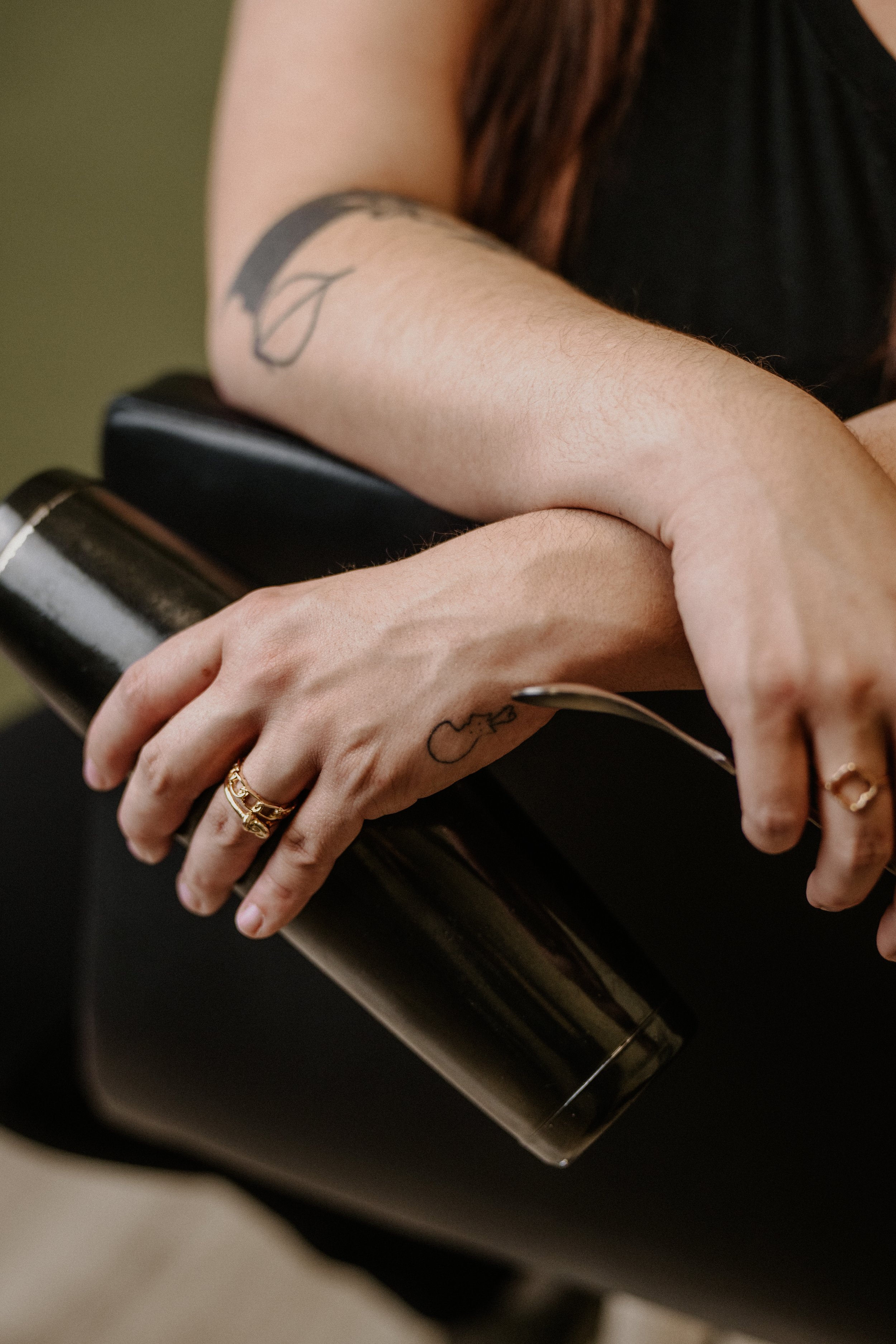 A person with a tattoo on their forearm, wearing multiple rings, holding a stainless steel cocktail mixer with a stirring spoon.