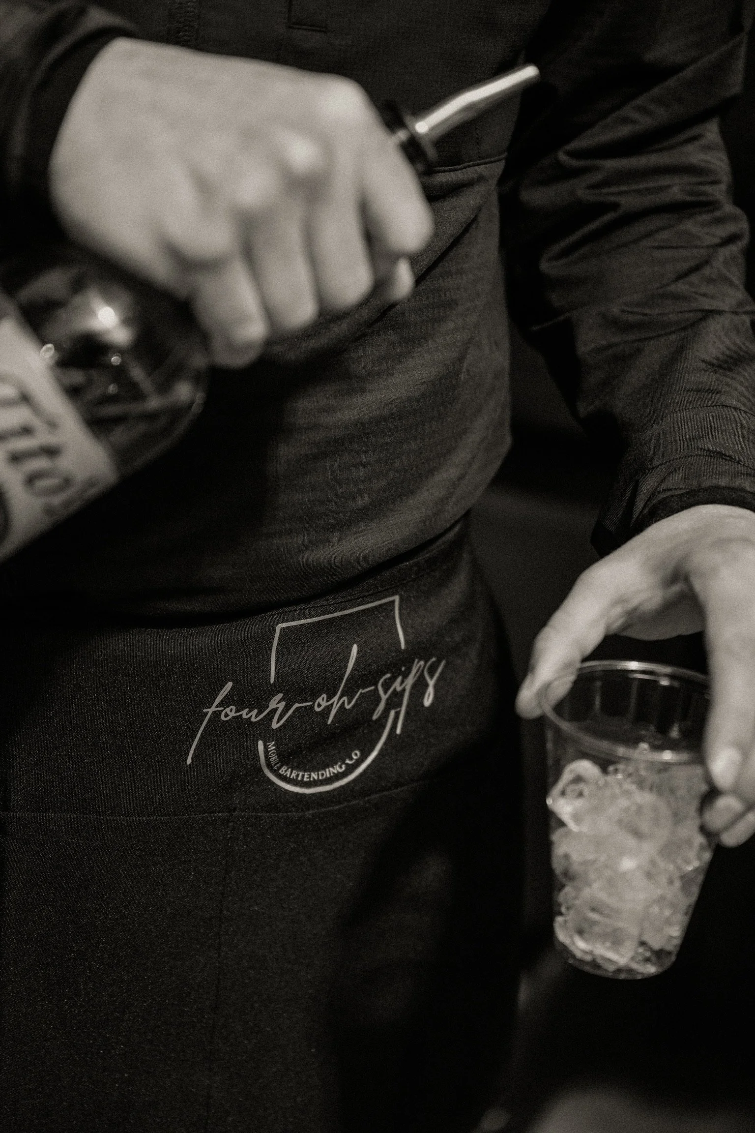 Close-up of a bartender's hand pouring a drink from a bottle into a glass, wearing a black apron with white text and Four Oh Sips logo, in a dark setting.