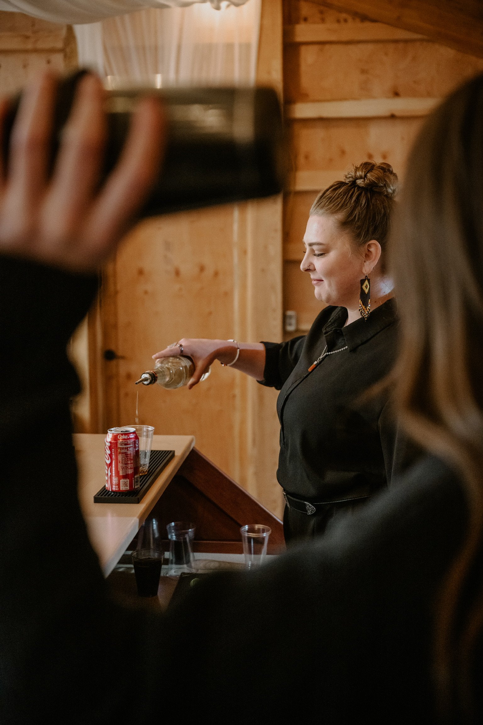 A woman pouring a drink from a bottle at a wooden bar with a Coca-Cola can and glasses on the counter, while a person in the foreground holds a phone.