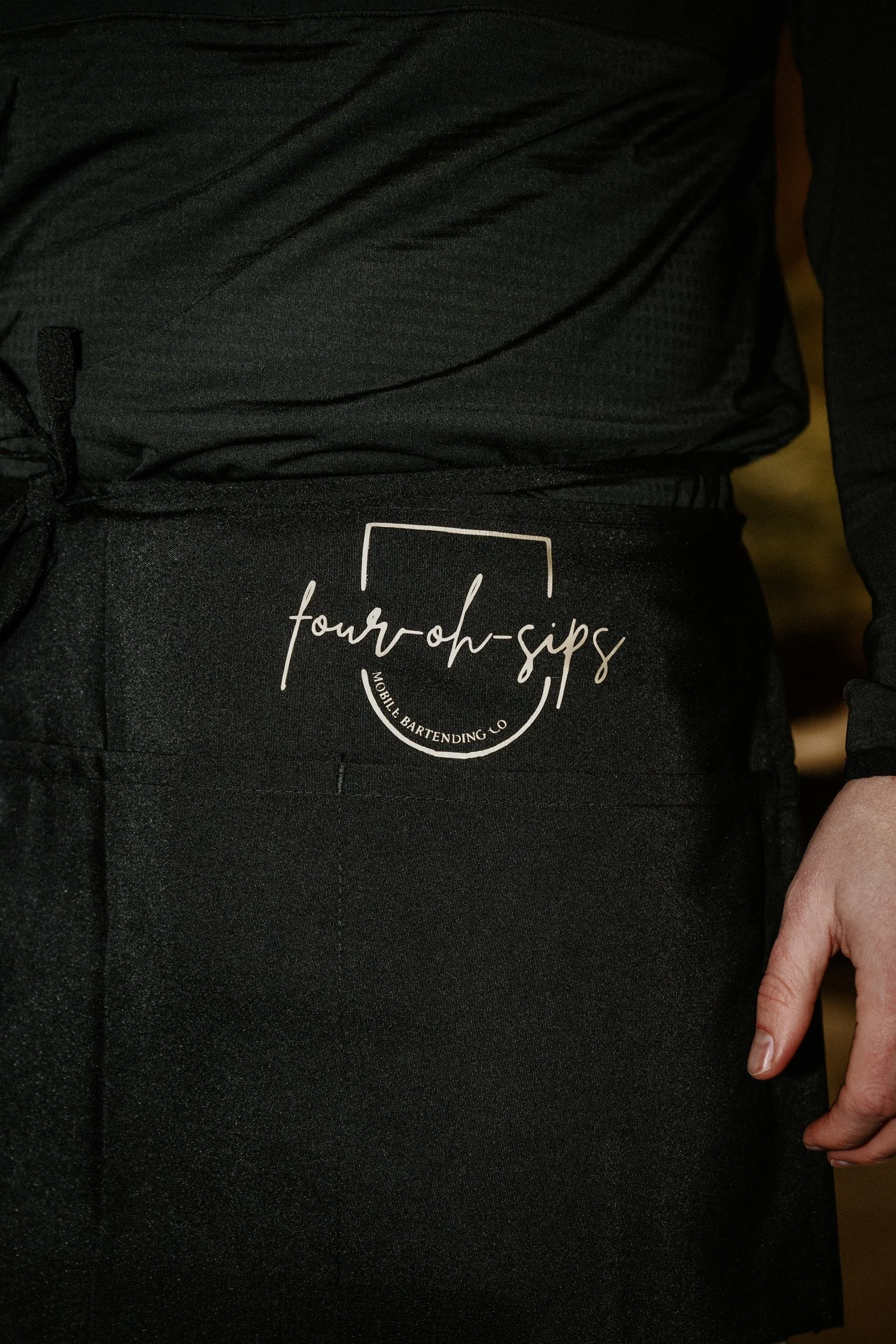 Close-up of person wearing black pants with a logo reading 'four-oh-sips' and 'Mobile Bartending Co.' printed on the front, with part of their hand visible.