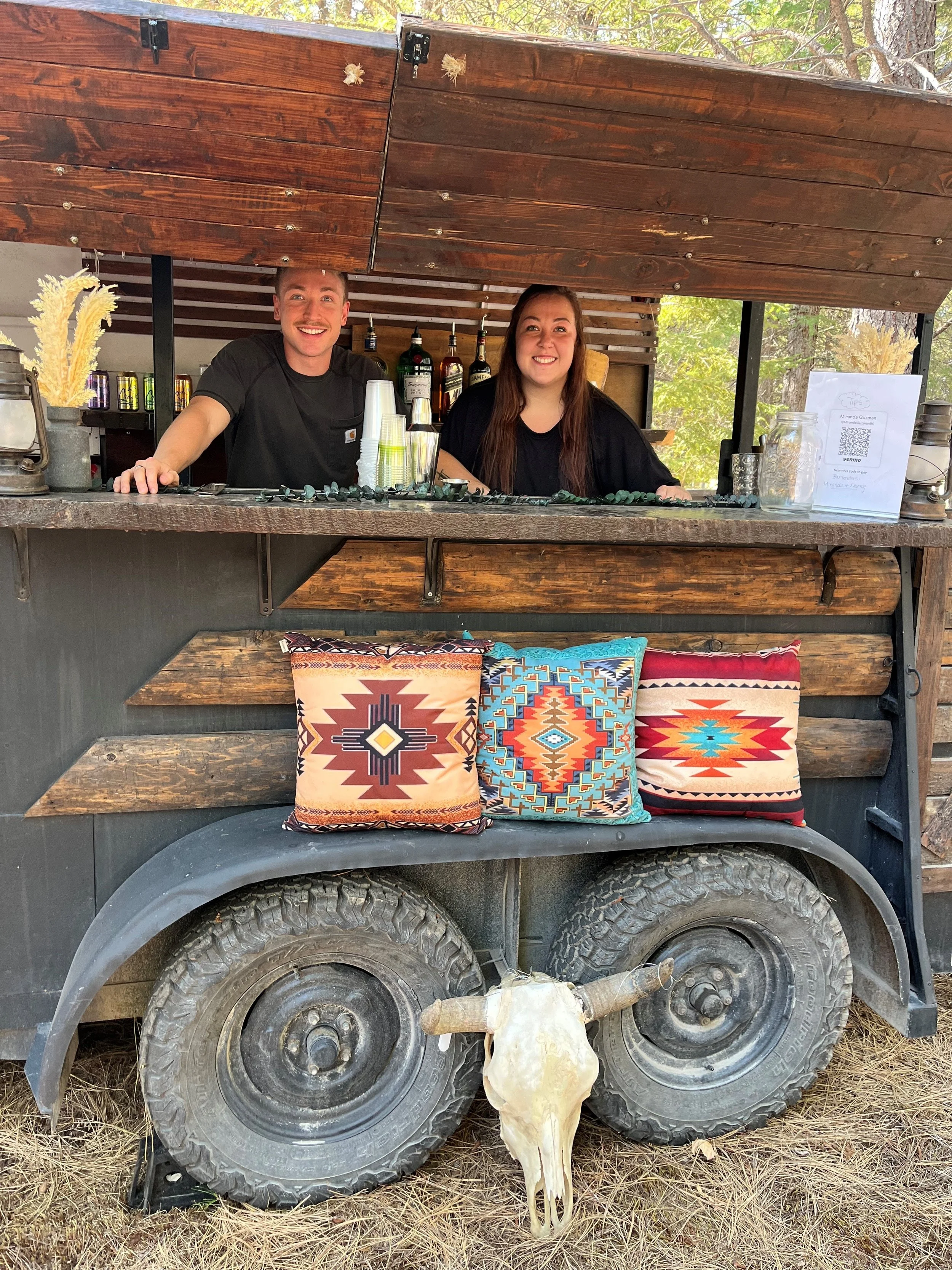 Two smiling people stand in a rustic outdoor bar cart with wooden panels, colorful decorative pillows, and a vintage skull with horns mounted on the front wheel. There are bottles and cups on the bar, and trees are visible in the background.