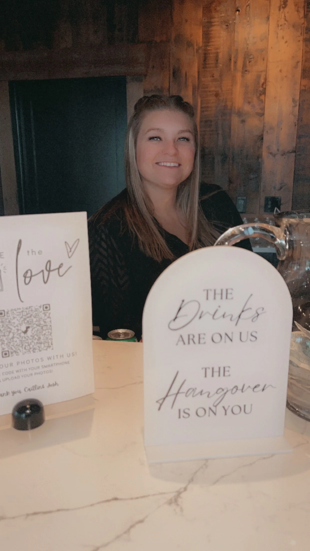 A smiling woman sitting at a table with signs that say "The drinks are on us, the hangover is on you" and a QR code for photos.