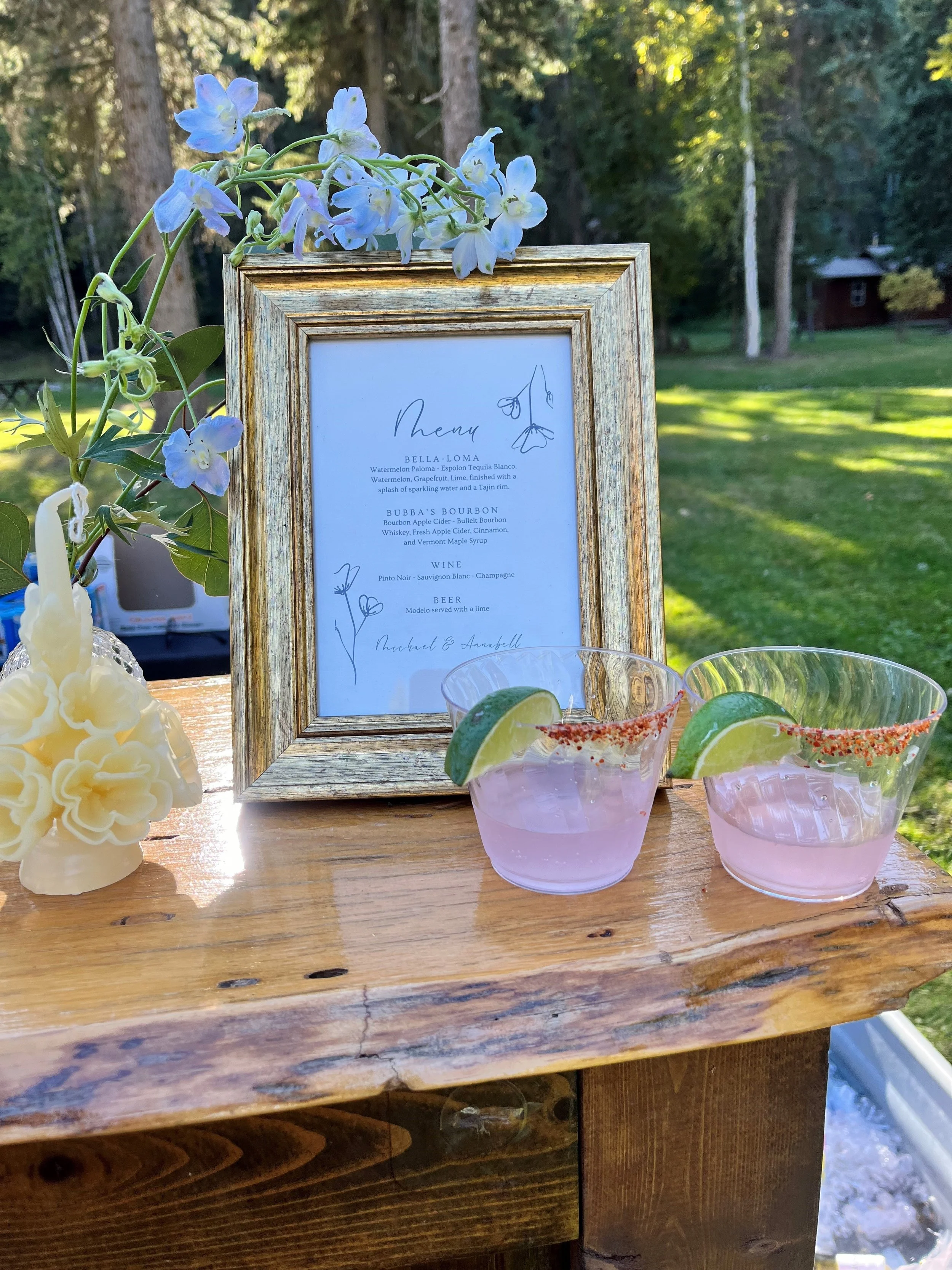 Outdoor table with two glasses of pink cocktail garnished with lime wedges and chili spice, a gold-framed menu with floral sketches, a flower arrangement, and a scenic background of trees and grass.