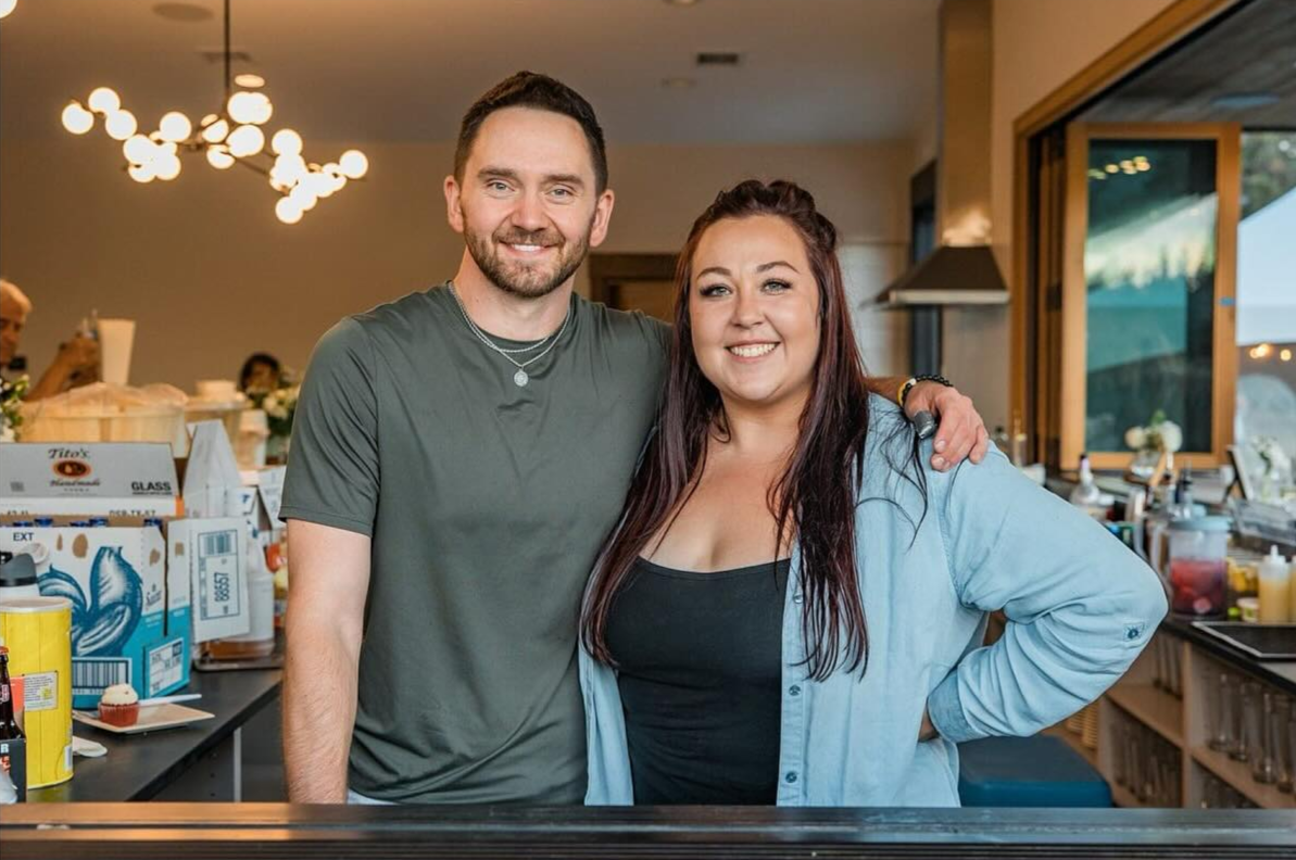 A smiling man and woman standing together in a kitchen, with the man having his arm around the woman. The background shows a kitchen with light fixtures and various kitchen supplies on the counter.